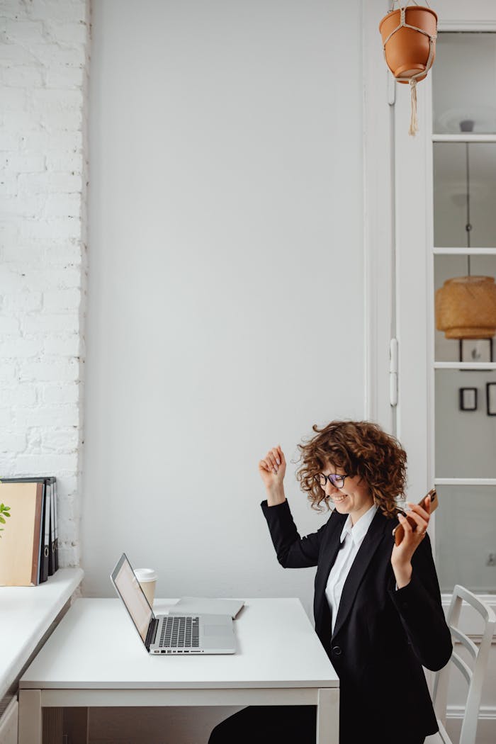 Home Businesswoman with curly hair celebrates success on phone call in modern office with laptop.